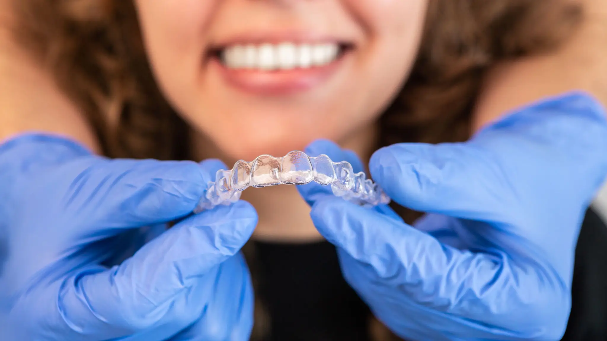 Person holding a clear dental aligner with blue gloves and smiling.